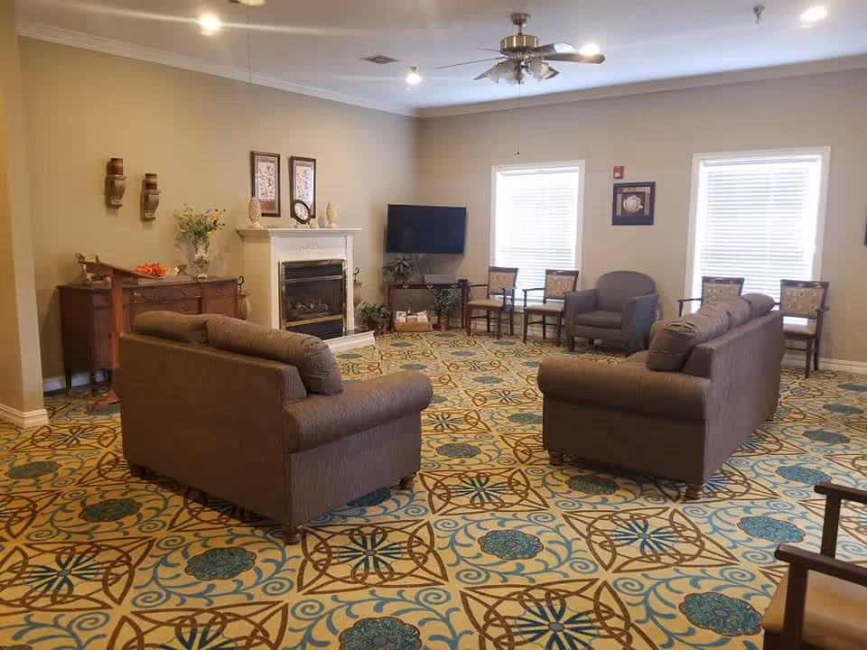 A spacious living room with two brown sofas facing each other, a fireplace with decorative items on the mantel, a flat-screen TV mounted on the wall, several chairs arranged along the walls, and a patterned carpet with blue and brown designs. The room has beige walls, two windows with blinds, and a ceiling fan with lights.