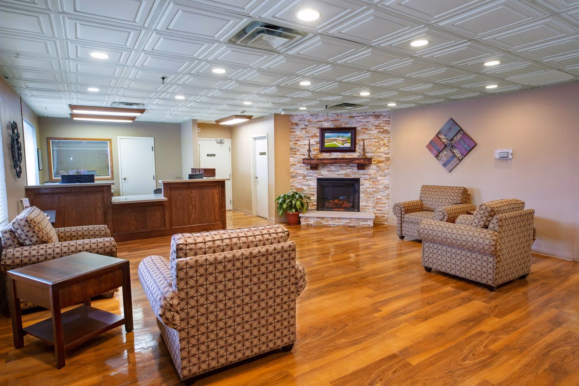 A spacious senior living facility common area with wooden flooring, patterned armchairs arranged around a stone fireplace with a framed picture above it, a wooden reception desk with computer monitors, and neutral-colored walls decorated with wall art and a potted plant.