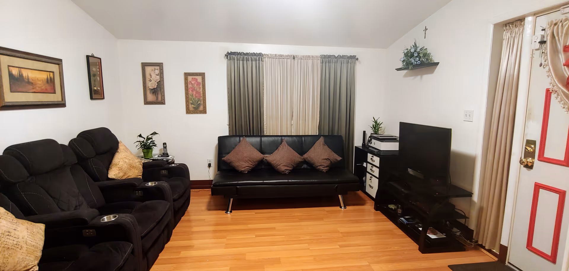 A cozy living room with wooden flooring, featuring a black leather sofa with three brown cushions, two black recliner chairs with beige pillows, a TV on a black stand, and a window with beige and green curtains. The walls are decorated with framed pictures and a small shelf with a plant, and there is a door with red trim on the right side.
