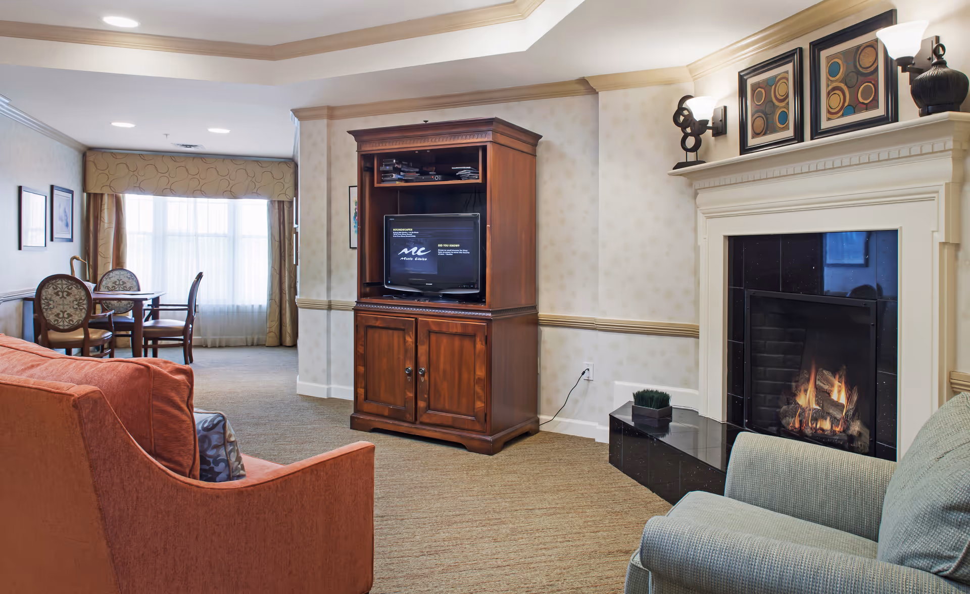 Communal living room with armchairs and a sofa facing a wooden TV cabinet and a lit fireplace, with a dining table by a window in the background.
