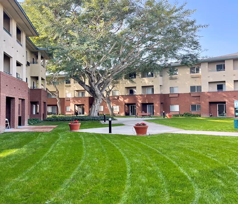Outdoor courtyard area of Carmel Village Retirement Community with well-maintained green grass, large trees, potted plants with flowers, benches, and a multi-story building with balconies and windows surrounding the courtyard.