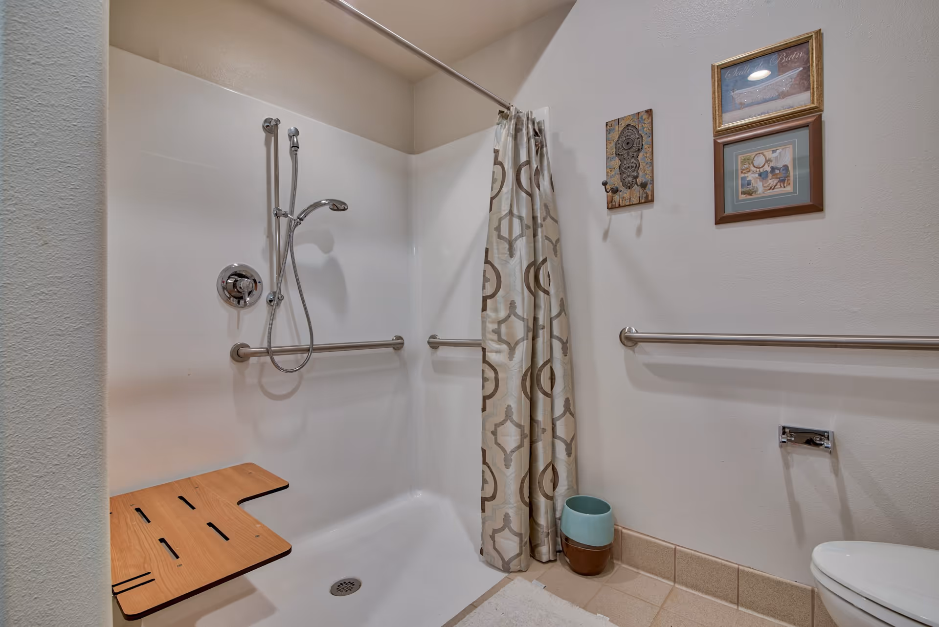 Accessible shower area with a foldable wooden seat, handheld showerhead, and grab bars on the walls. A patterned shower curtain is partially drawn to the side. The bathroom walls have two framed pictures and a decorative hook. A toilet paper holder and a small waste bin are visible near the toilet.