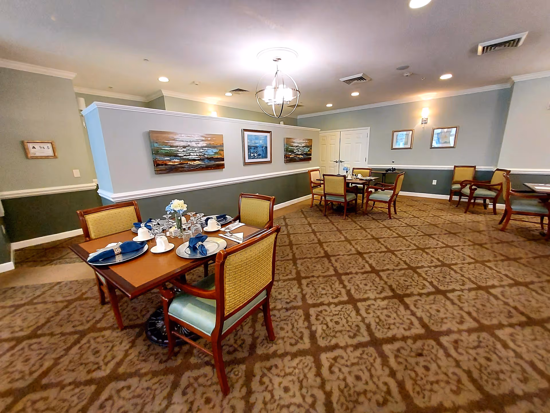 A dining room in a senior living facility with several wooden tables and chairs arranged neatly. Each table is set with plates, cups, napkins, and glassware. The room has patterned carpet flooring, light blue and green walls with white trim, and framed artwork hanging on the walls. Ceiling lights and a chandelier provide illumination.