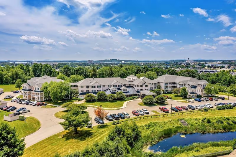 Aerial view of a large senior living facility named Solstice Senior Living at Bangor, surrounded by greenery, parking lots with cars, and a small pond in the foreground under a partly cloudy blue sky.