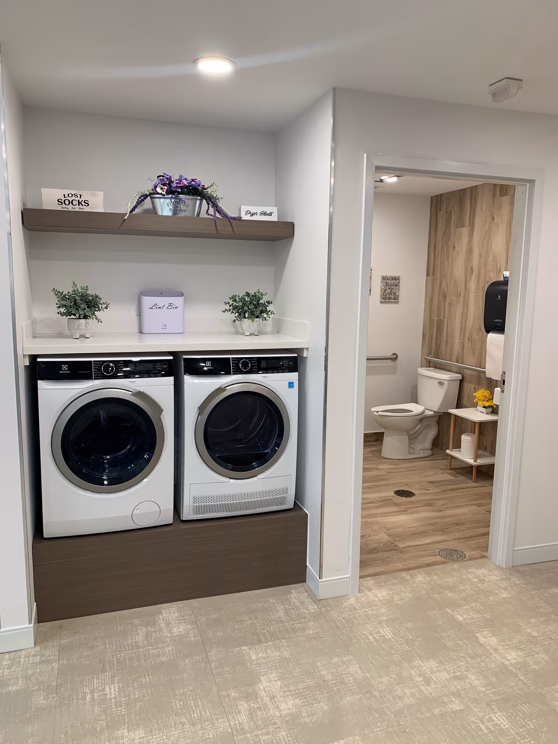 Laundry area with a front-loading washing machine and dryer side by side under a white countertop. Above the machines is a wooden shelf with a metal bucket of purple flowers and two small signs labeled 'Lost Socks' and 'Dryer Sheets'. To the right, an open doorway leads to a bathroom with a toilet, a small white shelf with yellow flowers, and wood-textured wall panels.