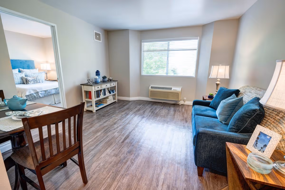 A bright living room with a blue sofa adorned with multiple cushions, a wooden side table with a lamp and picture frame, a small dining table with a chair, and a window with blinds letting in natural light. In the background, a bedroom with a bed, pillows, and a lamp is visible through an open doorway.