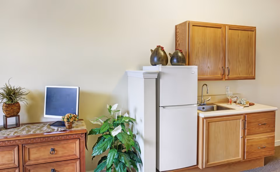 A small kitchenette area with a white refrigerator, wooden cabinets, a sink, and countertop. There are decorative items on top of the refrigerator and a wooden dresser with plants and a small chalkboard on it next to the kitchenette.