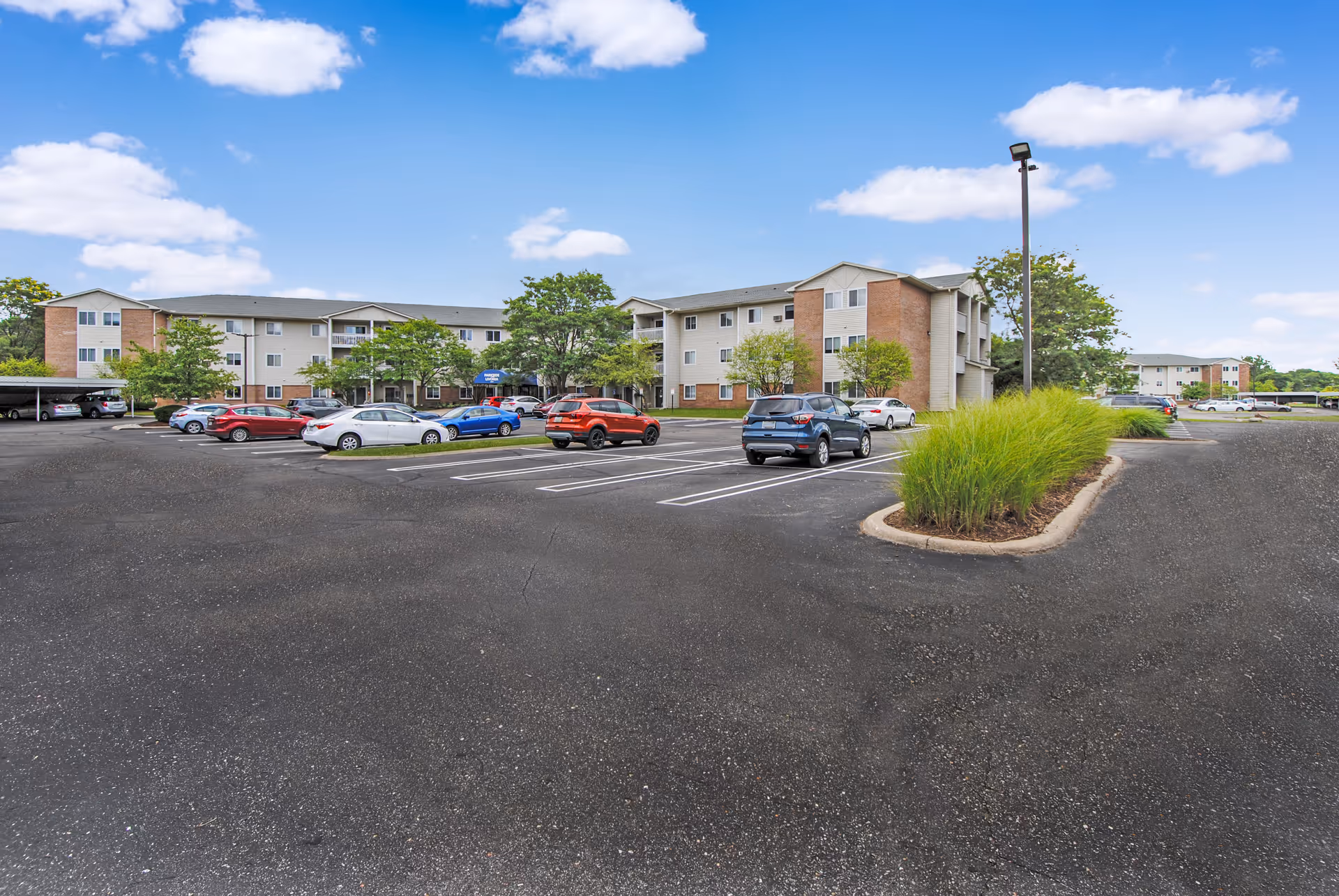 Exterior view of Parkside of Livonia senior living facility showing a large parking lot with several parked cars, landscaped areas with green bushes, and a three-story building under a blue sky with scattered clouds.