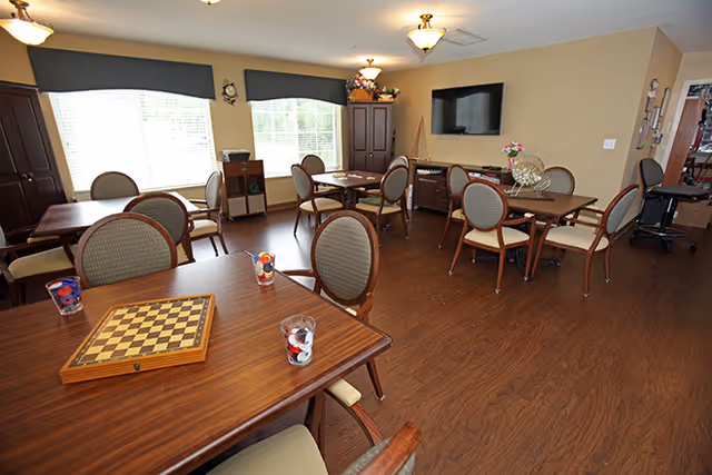 A well-lit common room with multiple wooden tables and cushioned chairs arranged neatly. One table has a chessboard and game pieces on it. The room features large windows with dark valances, a wall-mounted flat-screen TV, a cabinet, and decorative flowers. The floor is wooden, and the walls are painted beige.