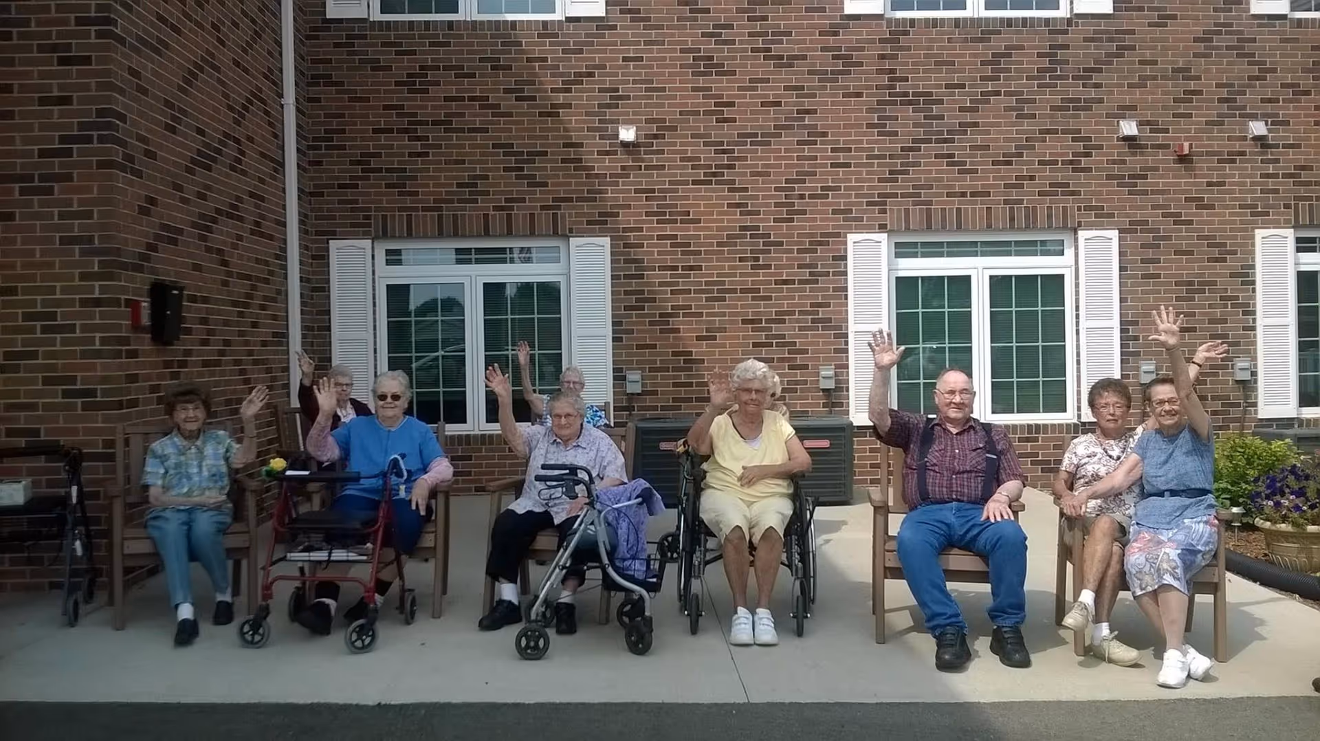 A group of elderly people sitting outside in front of a brick building, some in wheelchairs and others on chairs, all waving and smiling at the camera.