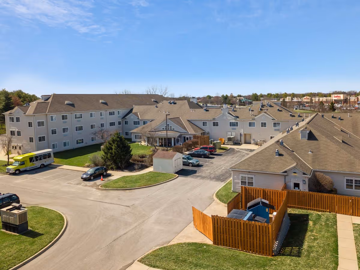 Aerial view of a multi-building senior living complex with parking areas, a fenced service enclosure, and a shuttle bus.