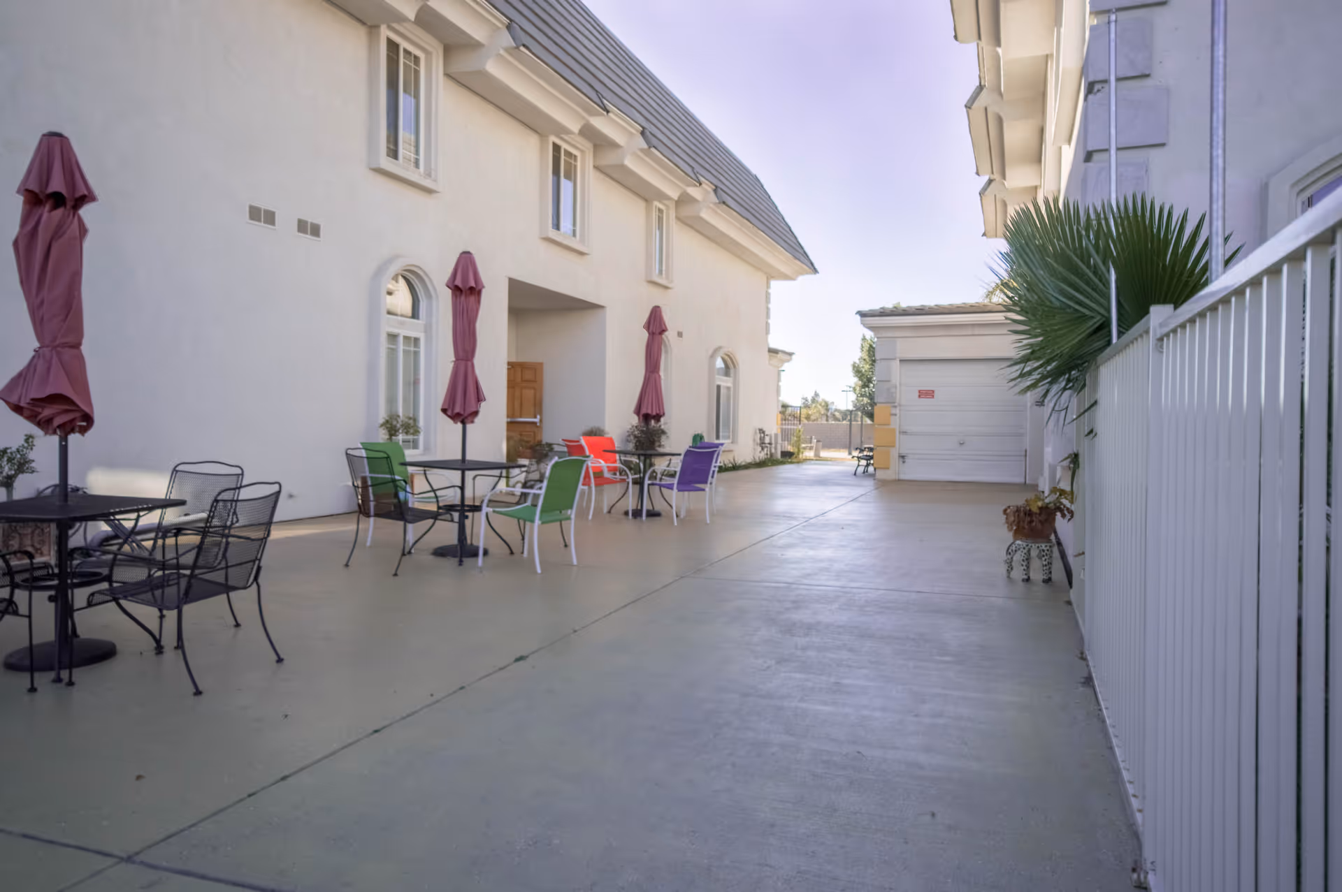 Outdoor patio area at Parkside Mansion Assisted Living Community with several tables and chairs, each table shaded by a closed umbrella. The patio is adjacent to a white building with multiple windows and a door, and there is a white fence on the right side with some potted plants.