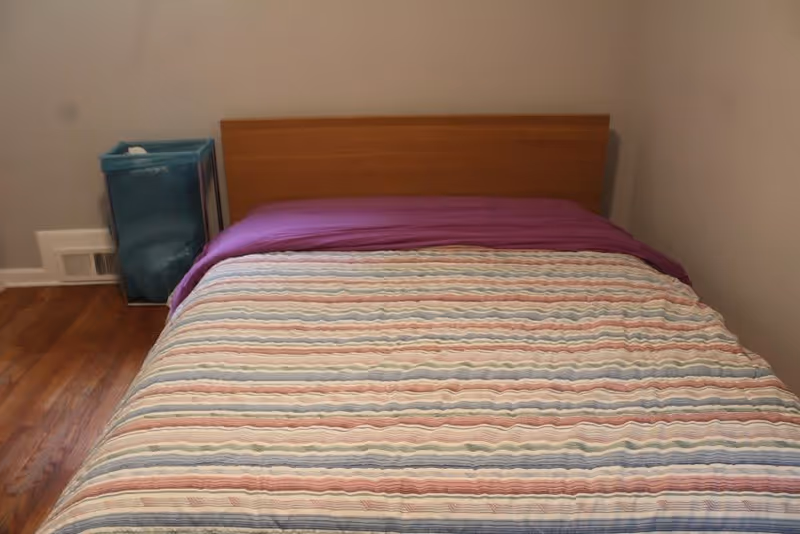 A simple bedroom with a wooden bed frame, a bed covered with a striped quilt in pastel colors, and a purple blanket folded at the head of the bed. There is a blue laundry hamper or basket placed on the wooden floor to the left side of the bed against a light-colored wall.