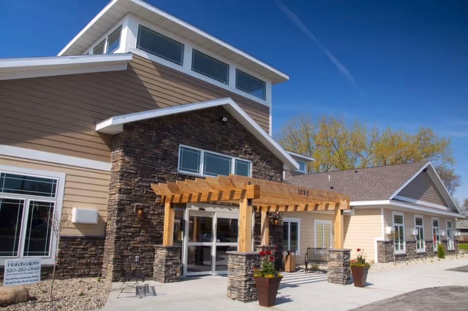 Front entrance of a modern senior living building with a stone facade, wooden pergola, planters, and a clear blue sky.