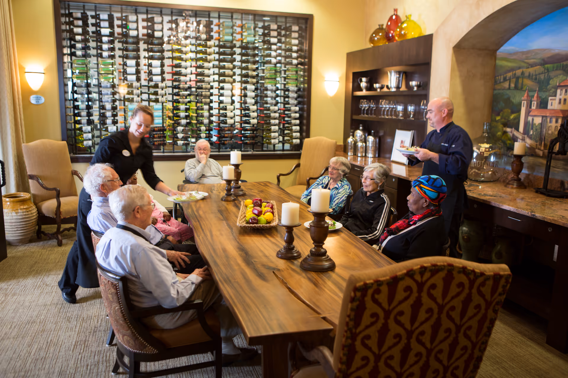 A group of elderly people sitting around a large wooden dining table in a warmly lit room with a wall-mounted wine rack. Two staff members are serving food to the residents. The room has comfortable chairs, decorative candles on the table, and shelves with glassware and decorative items.
