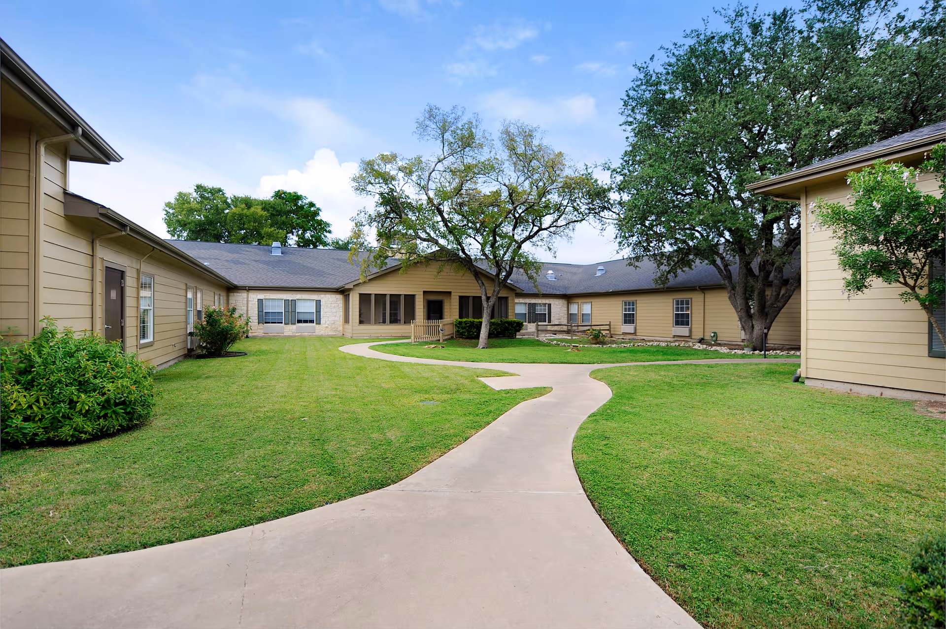 Courtyard with a curved concrete pathway, green lawn, trees, and single-story tan buildings under a blue sky.