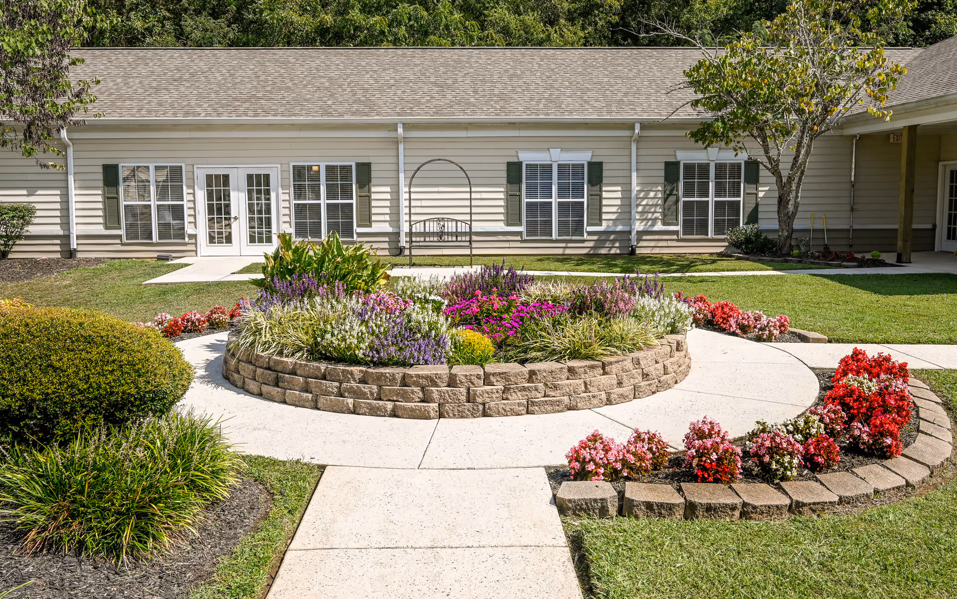 A landscaped outdoor garden area at Charter Senior Living of Cleveland featuring a circular raised flower bed with colorful flowers and greenery. Surrounding the flower bed is a curved concrete walkway. The building in the background has beige siding, multiple windows with green shutters, and a set of white double doors.