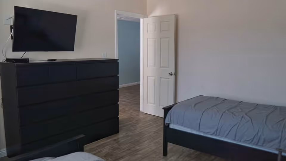 A simple bedroom with two single beds with gray bedding, a black dresser with multiple drawers, and a flat-screen TV mounted on the wall above the dresser. The room has light-colored walls and wood flooring, with a white door open to an adjacent room.