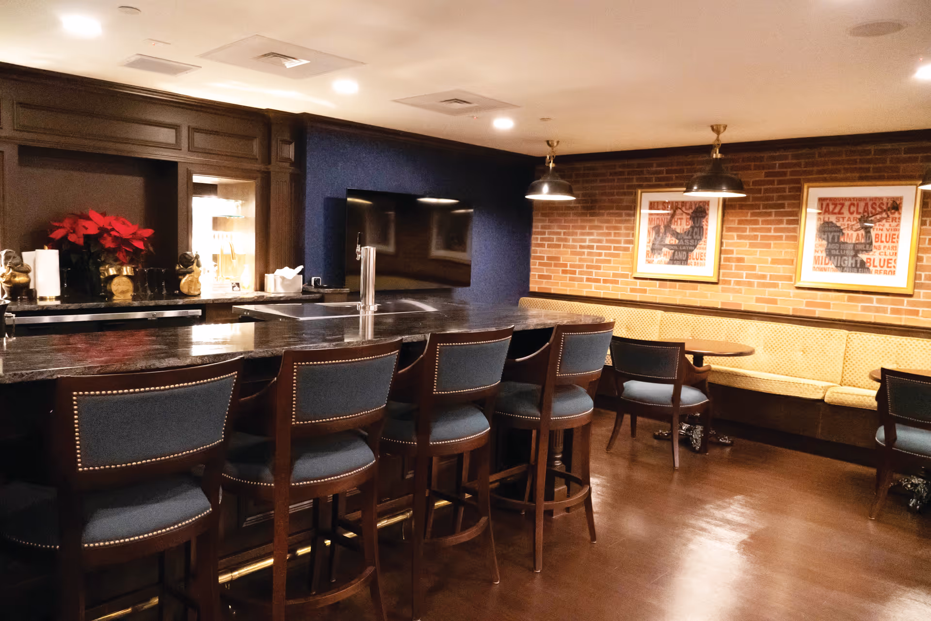 Interior lounge/bar with a long dark countertop, several upholstered bar stools, and banquette seating along a brick wall with framed art.