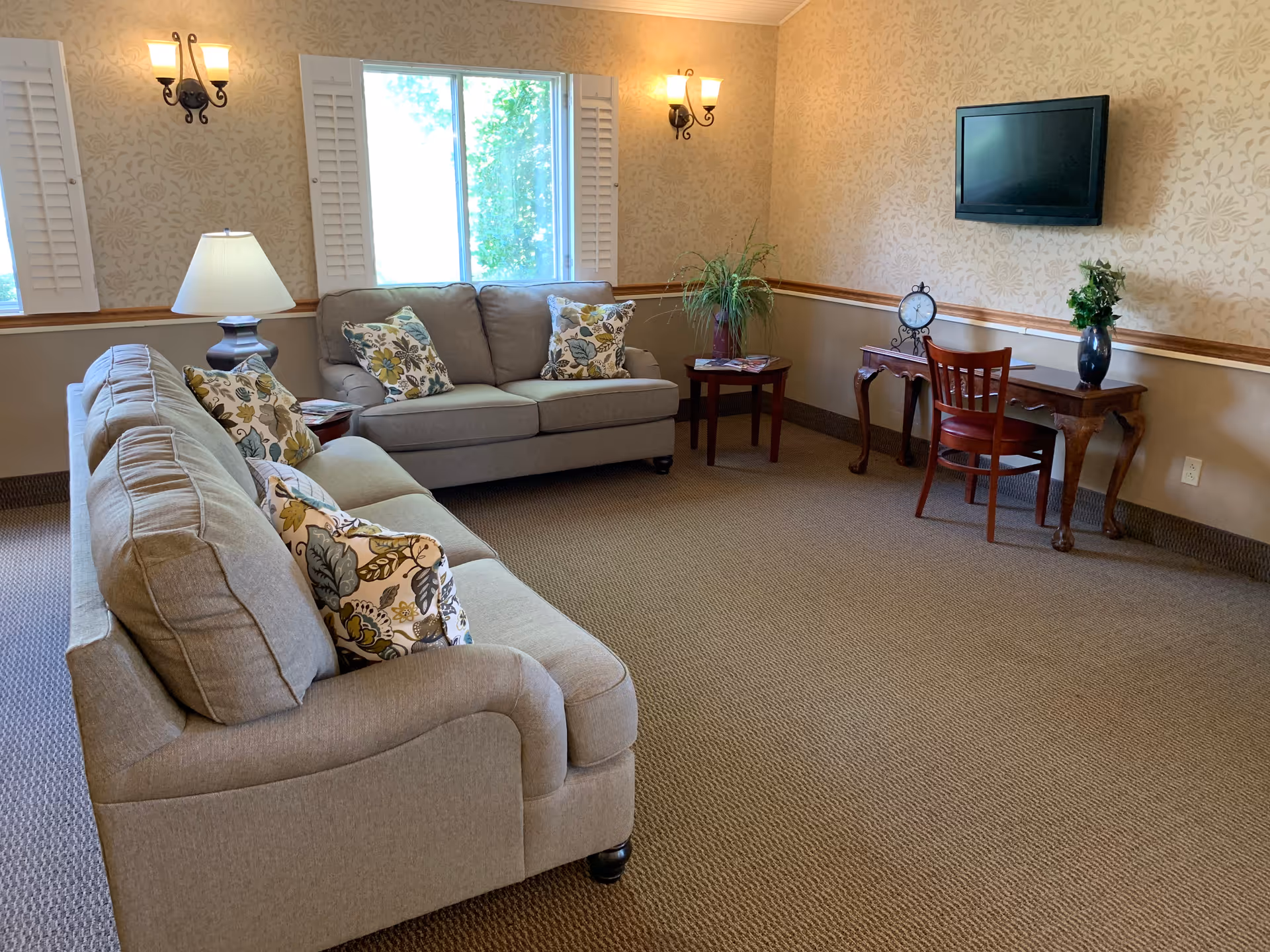 A cozy living room with two beige sofas adorned with floral patterned pillows, a window with white shutters letting in natural light, two wall-mounted sconces, a table lamp on a side table, a wooden desk with a chair, a clock, a vase with greenery, and a flat-screen TV mounted on the wall.
