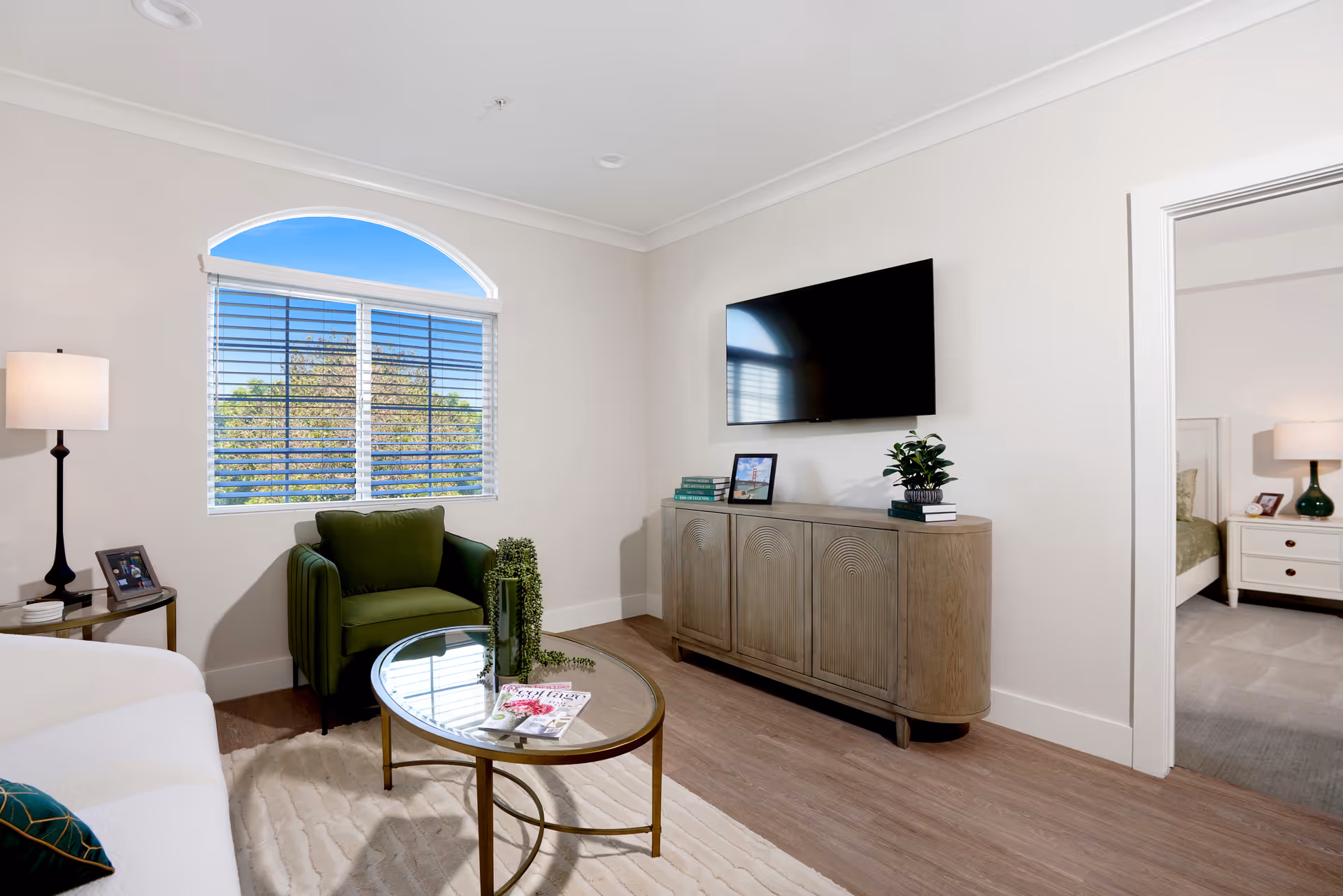 Bright living room with a wall-mounted TV, green armchair, round glass coffee table, and a wooden console cabinet.