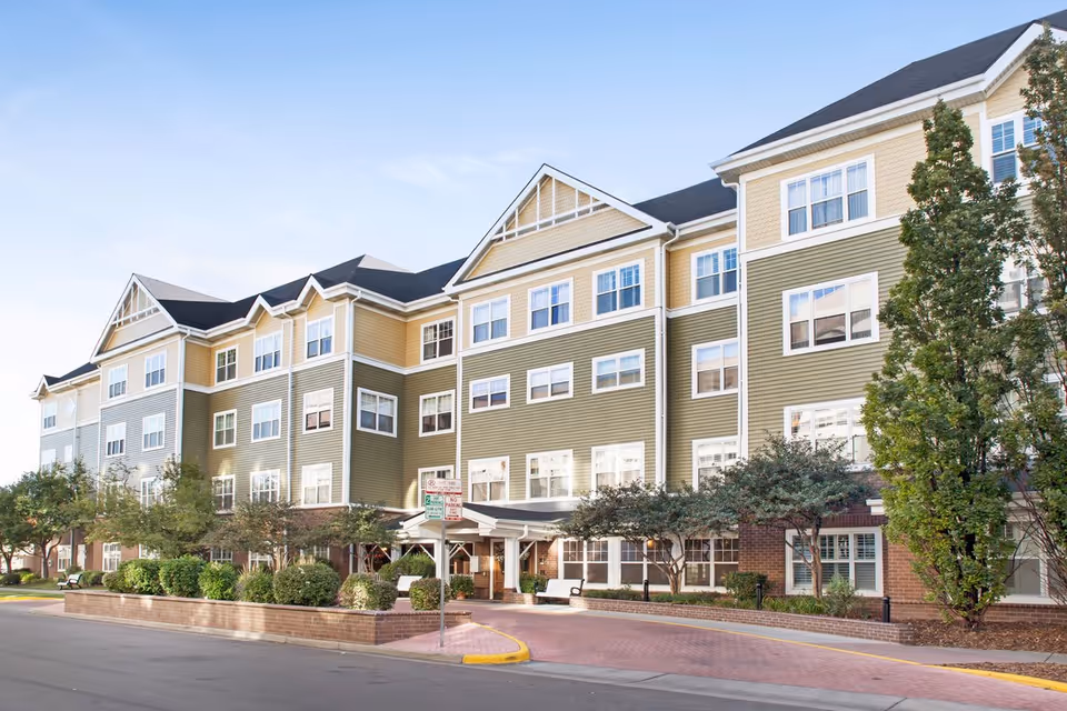 Front exterior of a multi-story senior living building with a covered entrance, many windows, and landscaped grounds.