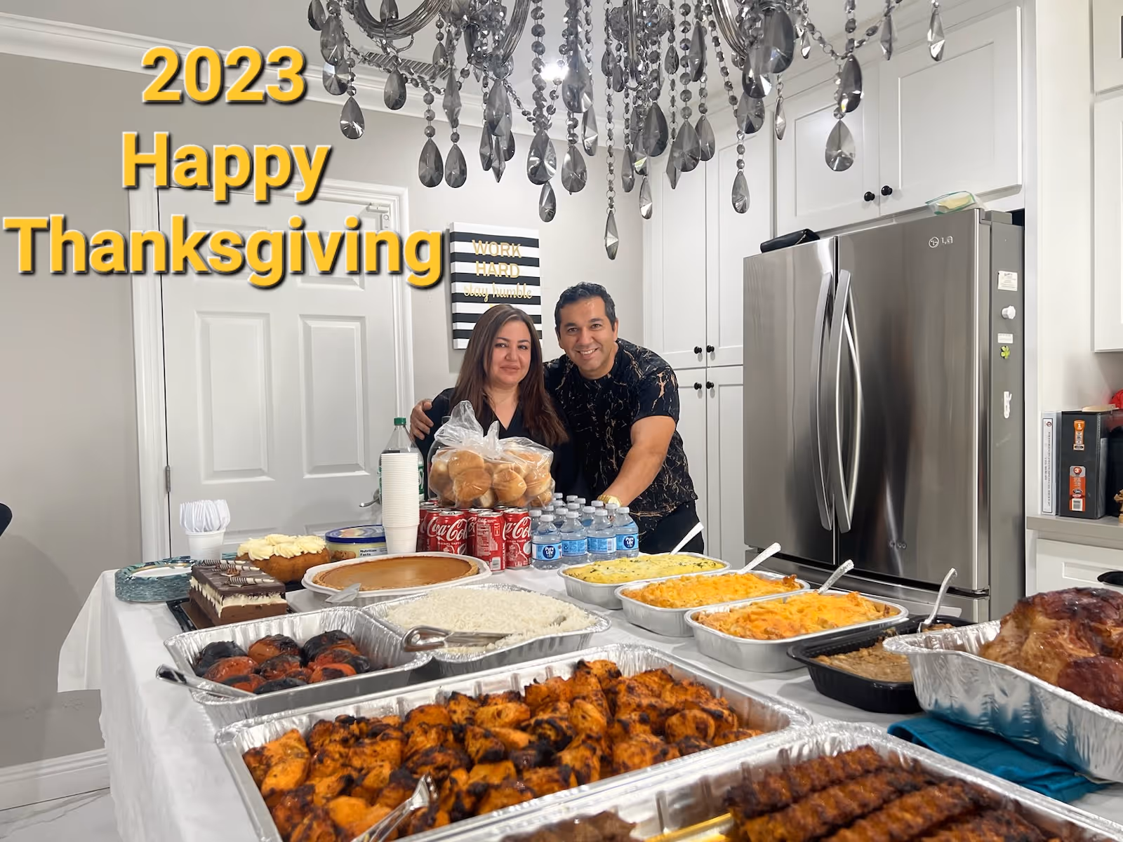 Two people stand behind a kitchen island piled with Thanksgiving dishes and drinks, with a stainless refrigerator and chandelier in the background.