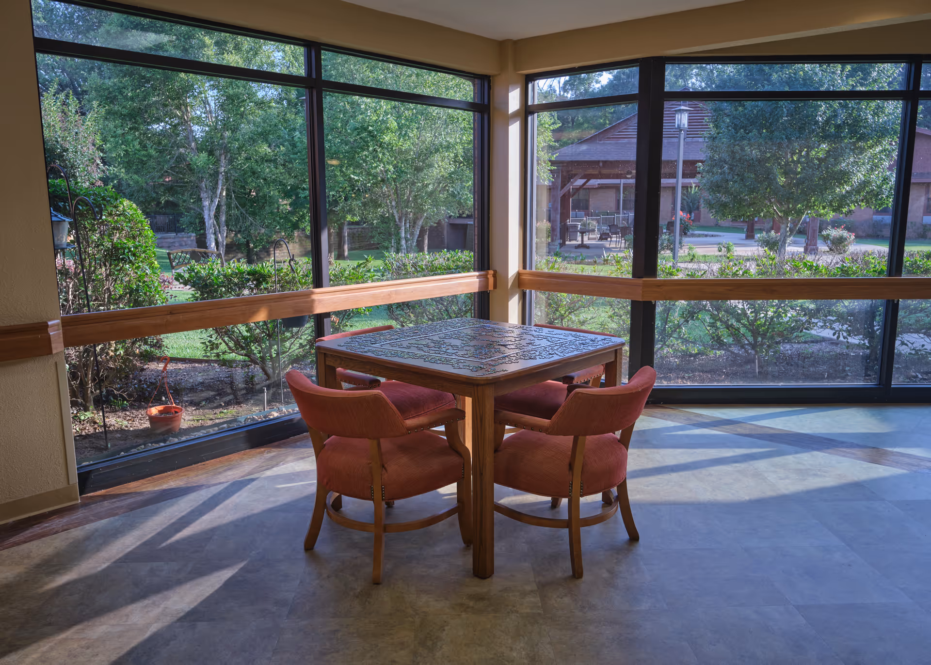 Sunlit seating area with a square puzzle table and four red upholstered chairs in front of large windows overlooking a garden.