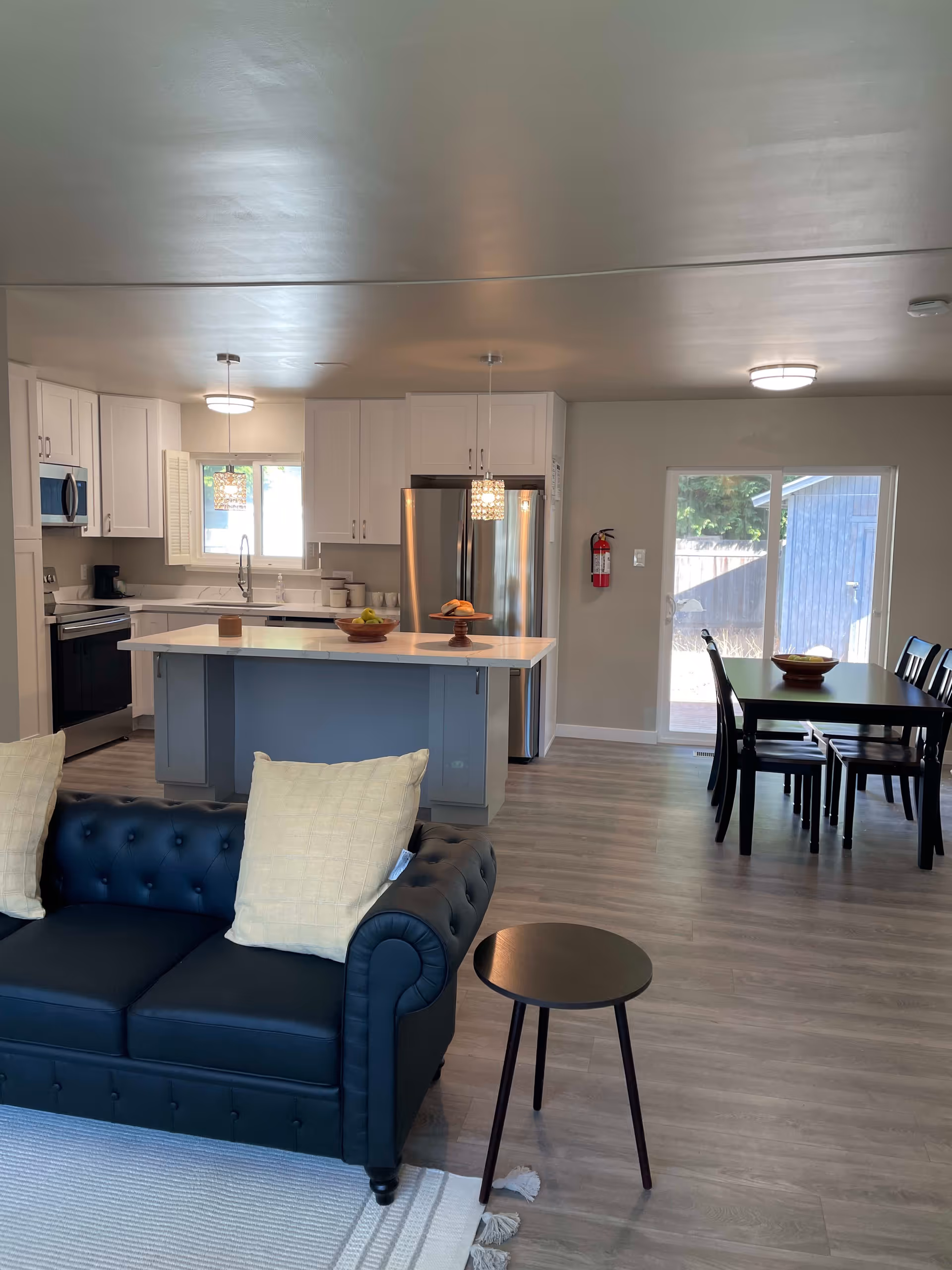 Open concept interior showing a modern kitchen with white cabinets, stainless steel appliances, and a central island with pendant lights. Adjacent to the kitchen is a dining area with a black table and chairs, and in the foreground is a black leather tufted sofa with two light-colored pillows and a small round black side table on a light rug.