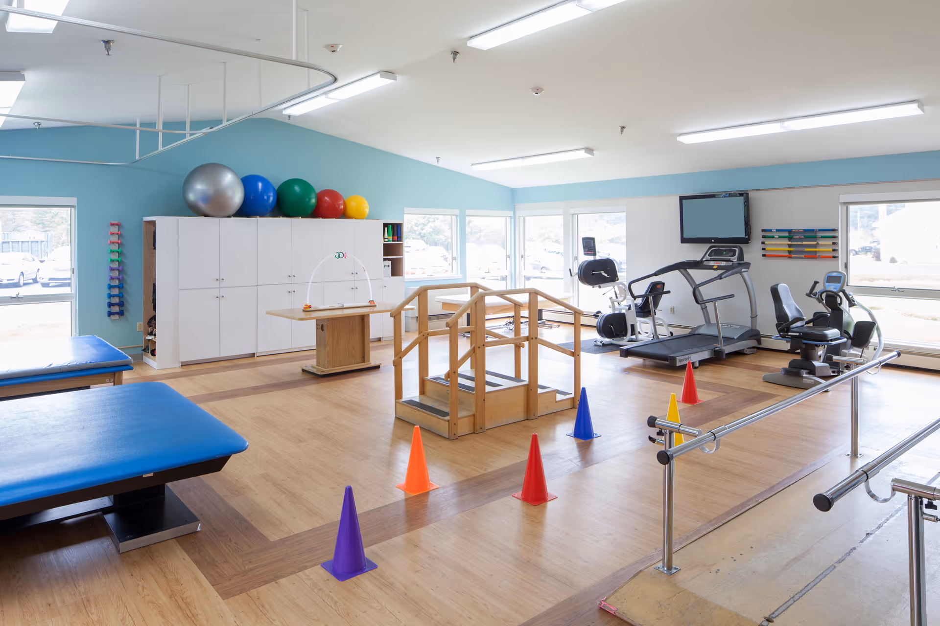 A bright physical therapy room with exercise equipment including a treadmill, stationary bikes, parallel bars, therapy tables, colorful exercise balls on top of cabinets, and small traffic cones arranged on the wooden floor. Large windows let in natural light.