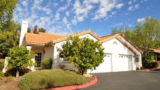 Single-story Mediterranean-style building with a tiled roof, attached two-car garage, and landscaped shrubs and trees under a blue sky.