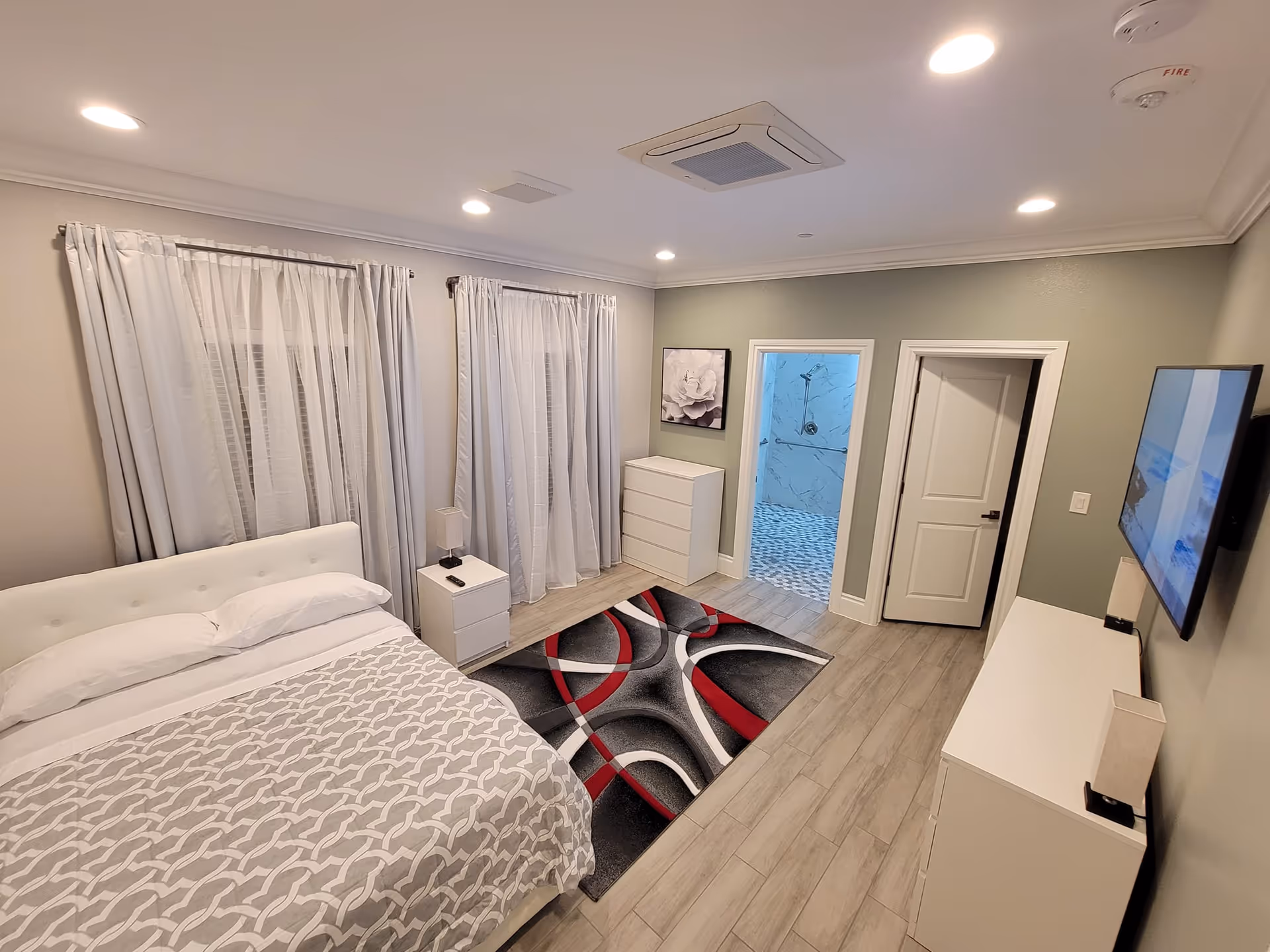 A modern bedroom with a white bed, nightstand with a lamp, two windows with white curtains, a dresser, a colorful area rug, and a wall-mounted TV. There are two doors, one leading to a bathroom with a walk-in shower and the other a closed door. The room has recessed lighting and light-colored wood flooring.
