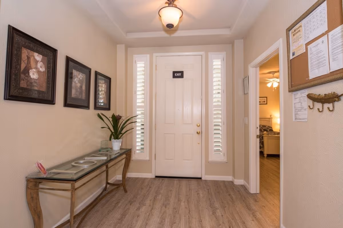 An entryway with a white door labeled 'EXIT' flanked by two narrow windows with white shutters. To the left, there is a glass-top console table with a potted plant and decorative items. Three framed pictures hang on the wall above the table. To the right, a doorway leads to a bedroom with a bed and ceiling fan visible. A bulletin board and coat hooks are mounted on the wall near the doorway. The floor is wood, and a ceiling light fixture is centered above.