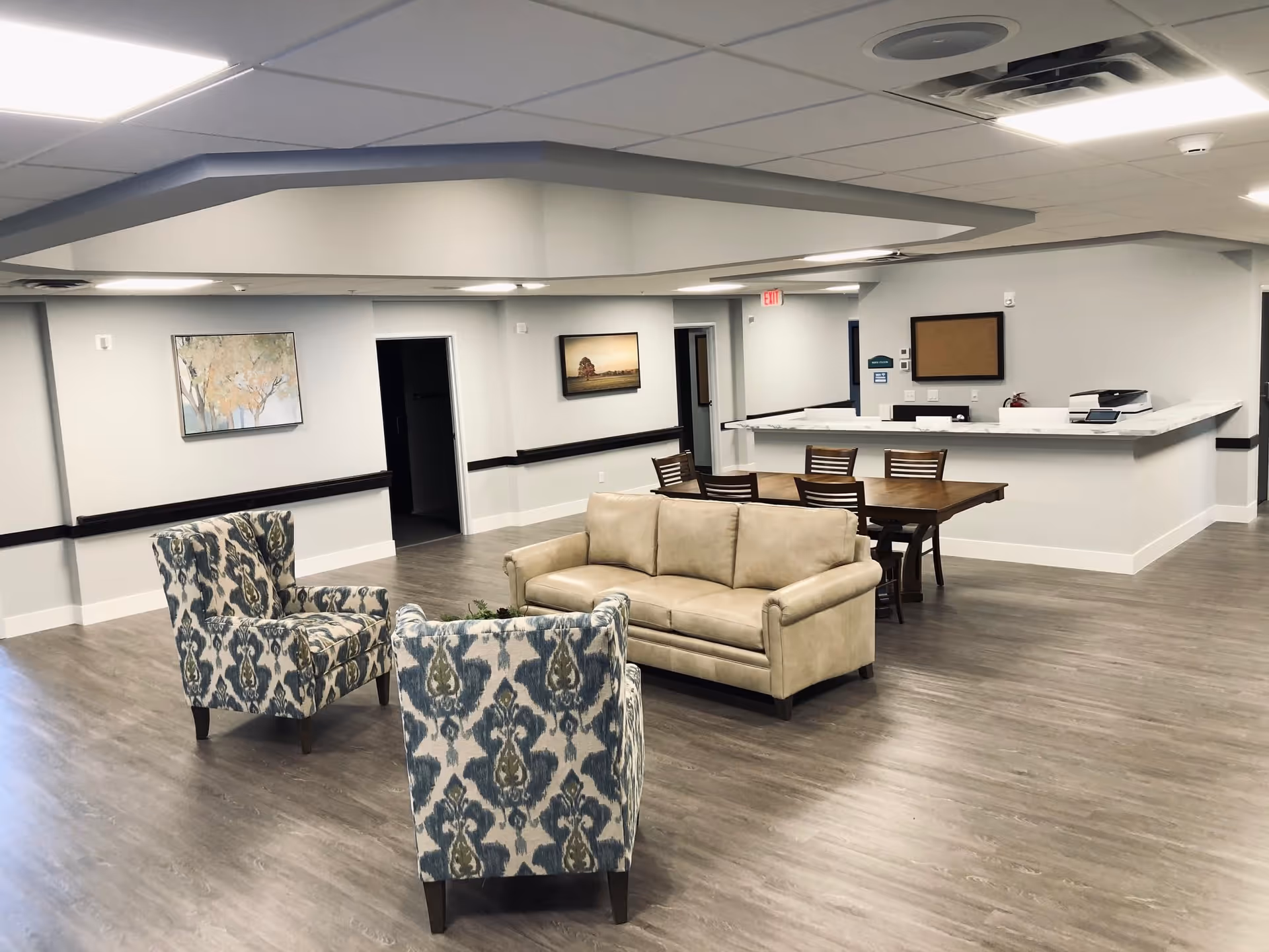 A spacious senior living facility common area with two patterned armchairs, a beige sofa, a wooden dining table with six chairs, and a reception desk with a marble countertop. The room has light gray walls, wood flooring, ceiling lights, and framed artwork on the walls.