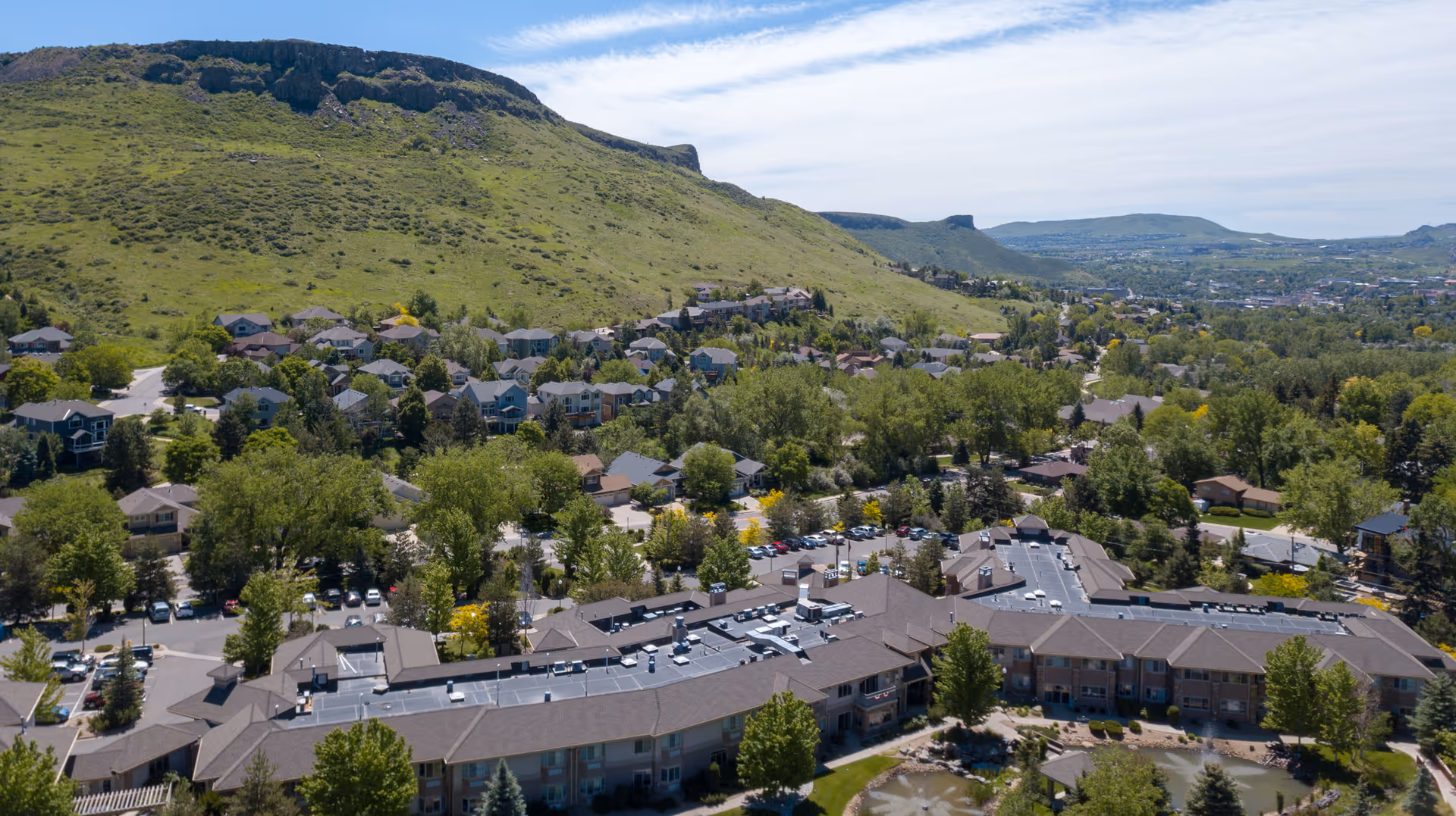 Aerial view of a senior living facility named Golden Pond Senior Living surrounded by trees and residential houses with a large green hill in the background under a partly cloudy sky.