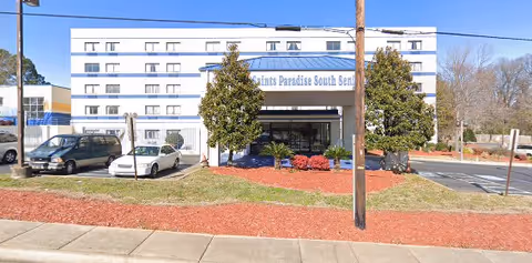 Front exterior view of a multi-story senior living facility named Madison Saints Paradise South with a parking area, trees, and landscaping in front.