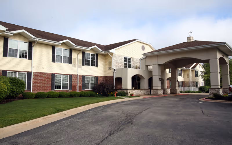 Exterior view of a two-story senior living facility building with a covered entrance driveway, beige siding, red brick lower facade, and multiple windows. The surrounding area includes a well-maintained lawn and shrubs under a partly cloudy sky.