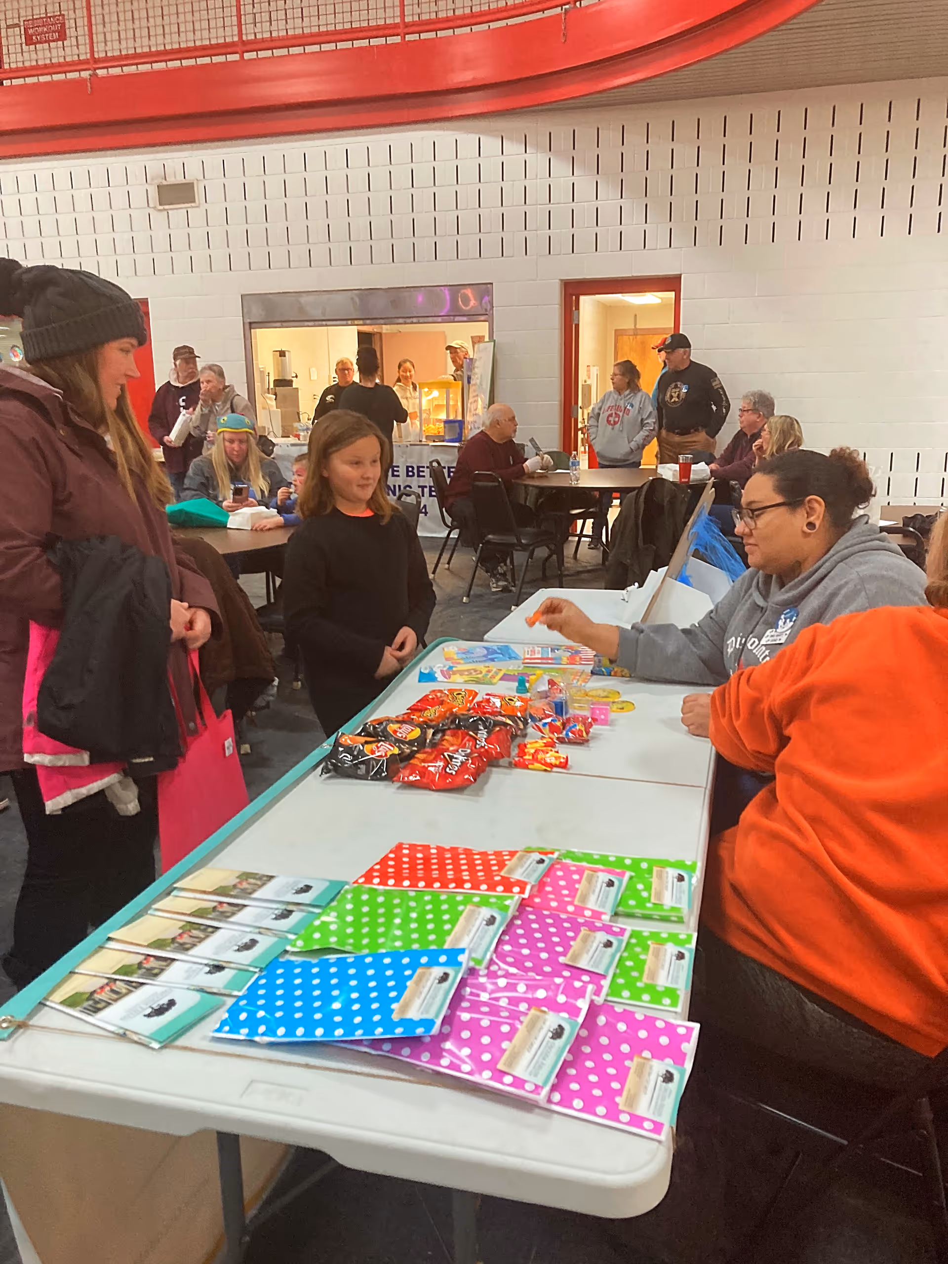 Indoor community event with people gathered around tables. A woman and a child stand on one side of a table covered with colorful polka dot gift bags and snacks, while two seated women on the other side engage with them. In the background, other people are seated at round tables and standing near a service window in a large room with white brick walls and a red railing above.