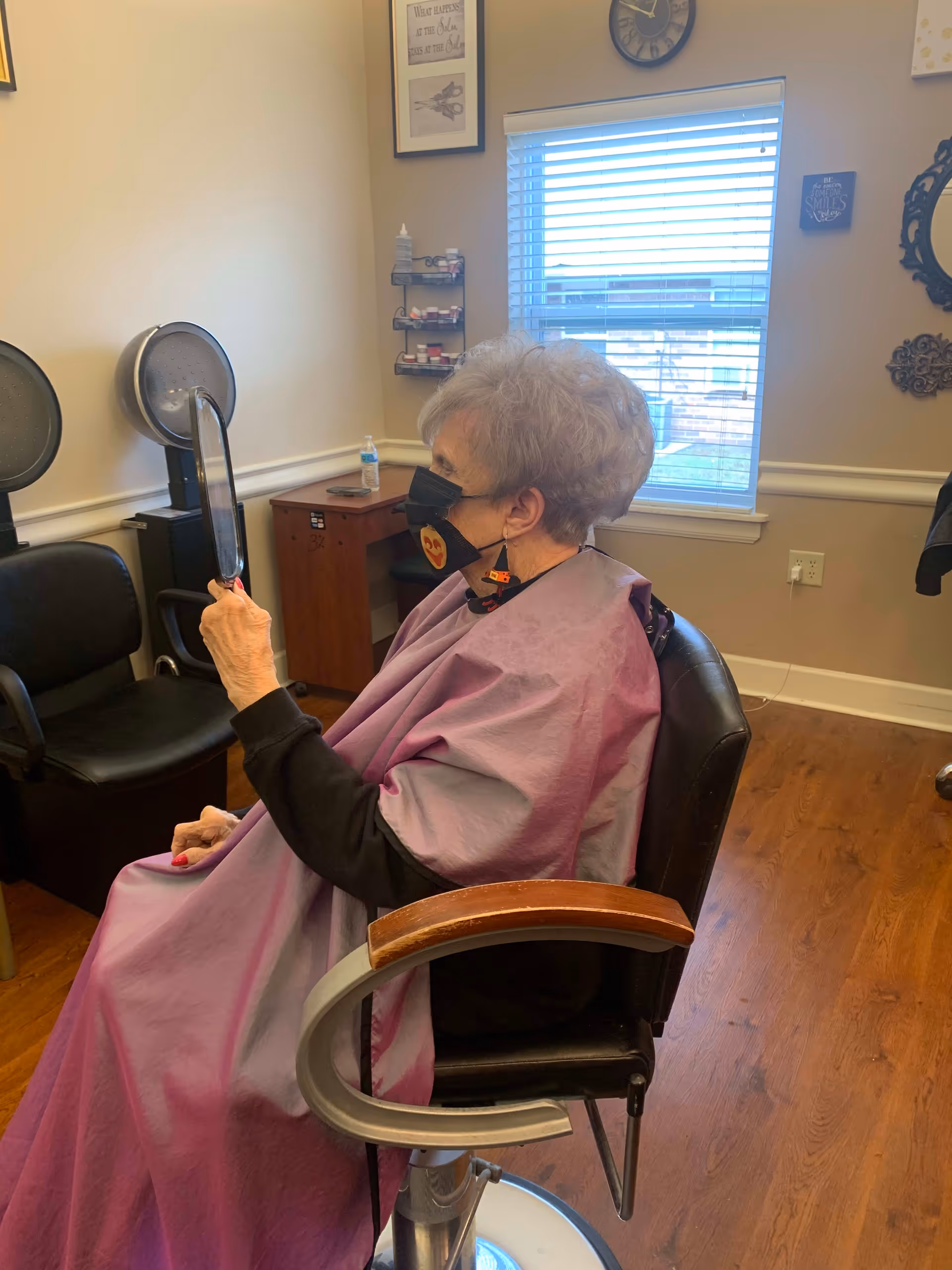 An elderly woman wearing a black face mask and a purple salon cape is sitting in a salon chair holding a hand mirror, looking at her reflection. The room has wooden flooring, a window with blinds, and salon equipment including hair dryers and chairs. Various wall decorations and a clock are visible.