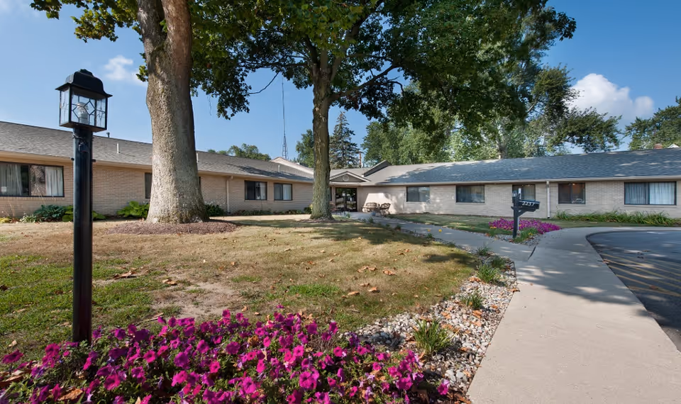 Single-story brick facility front with a paved walkway, large trees, a lamp post, and purple flowerbeds.