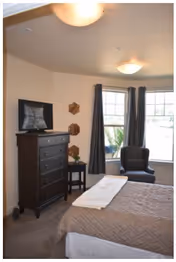 A bedroom with a bed covered in a brown quilt and a white towel folded on top. There is a dark wooden dresser with a flat-screen TV on it, a small side table with a plant, and a dark armchair near two large windows with dark curtains. The room has beige walls and carpeted floor.