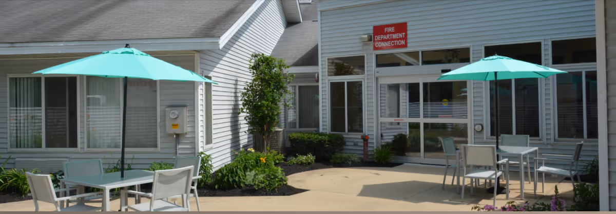 Outdoor patio area at Glendale Care and Rehab Center with two tables, each shaded by a turquoise umbrella, surrounded by chairs. The building exterior is light gray with multiple windows and a door. There is a red sign above the door that reads 'FIRE DEPARTMENT CONNECTION'.