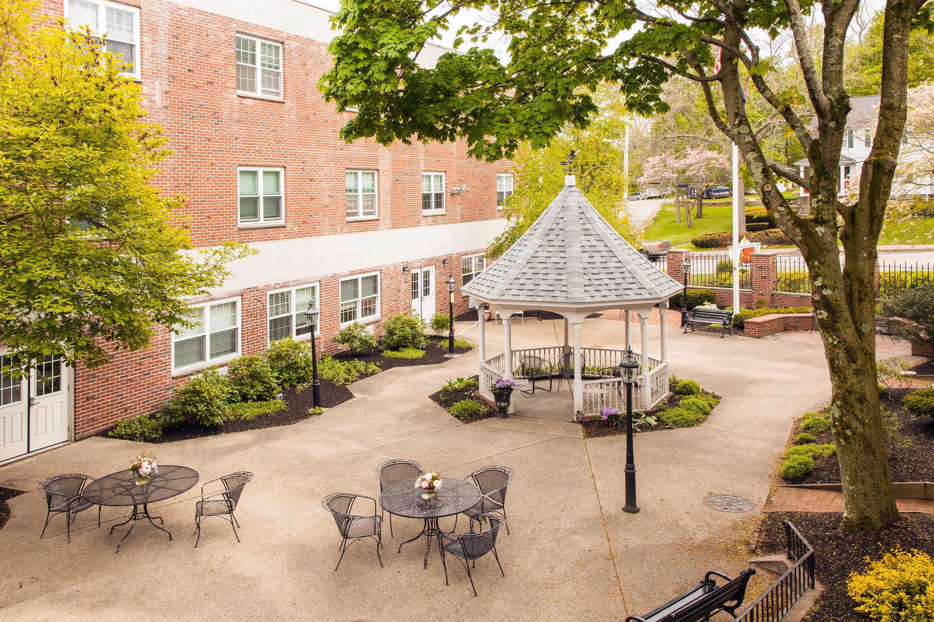 Courtyard of a brick senior living facility with a central white gazebo, patio tables, benches and surrounding trees.