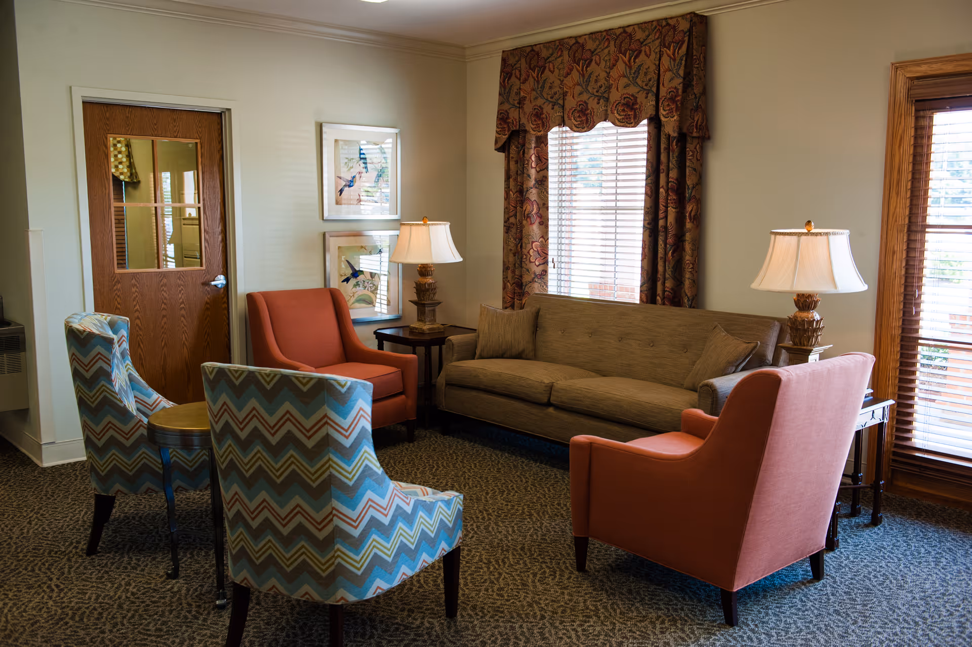 A cozy living room area with a beige sofa, two coral armchairs, and two patterned armchairs arranged around a small round table. There are two table lamps on side tables, framed bird artwork on the wall, and windows with wooden blinds and patterned valances.