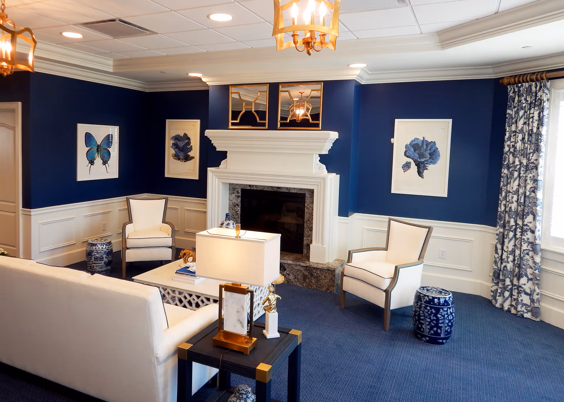 A cozy living room with navy blue walls and white wainscoting, featuring a white fireplace with a granite hearth. The room includes two white armchairs with dark trim, a white sofa, a white coffee table, and a side table with a lamp. Blue and white decorative ceramic stools and framed artwork of blue flowers and a butterfly adorn the walls. A window with blue and white patterned curtains lets in natural light.