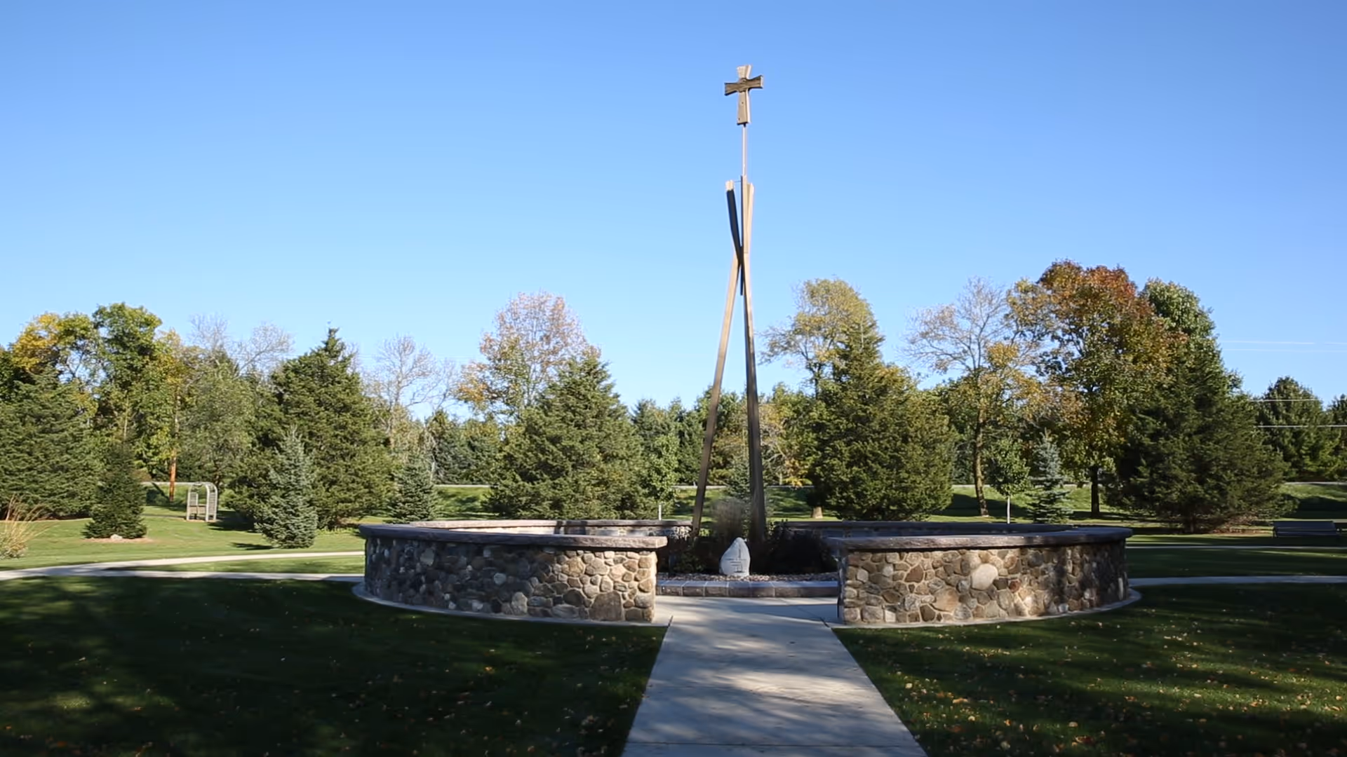 Outdoor area with a circular stone wall surrounding a tall wooden cross structure. The area is surrounded by green grass, trees, and a clear blue sky.