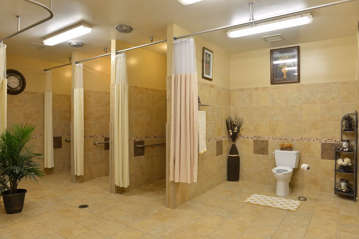 A spacious bathroom with beige tiled walls and floor, featuring three shower stalls with cream-colored curtains and a toilet area with a white toilet, a small rug, and decorative items including a potted plant, framed pictures, and a shelving unit with ornaments.