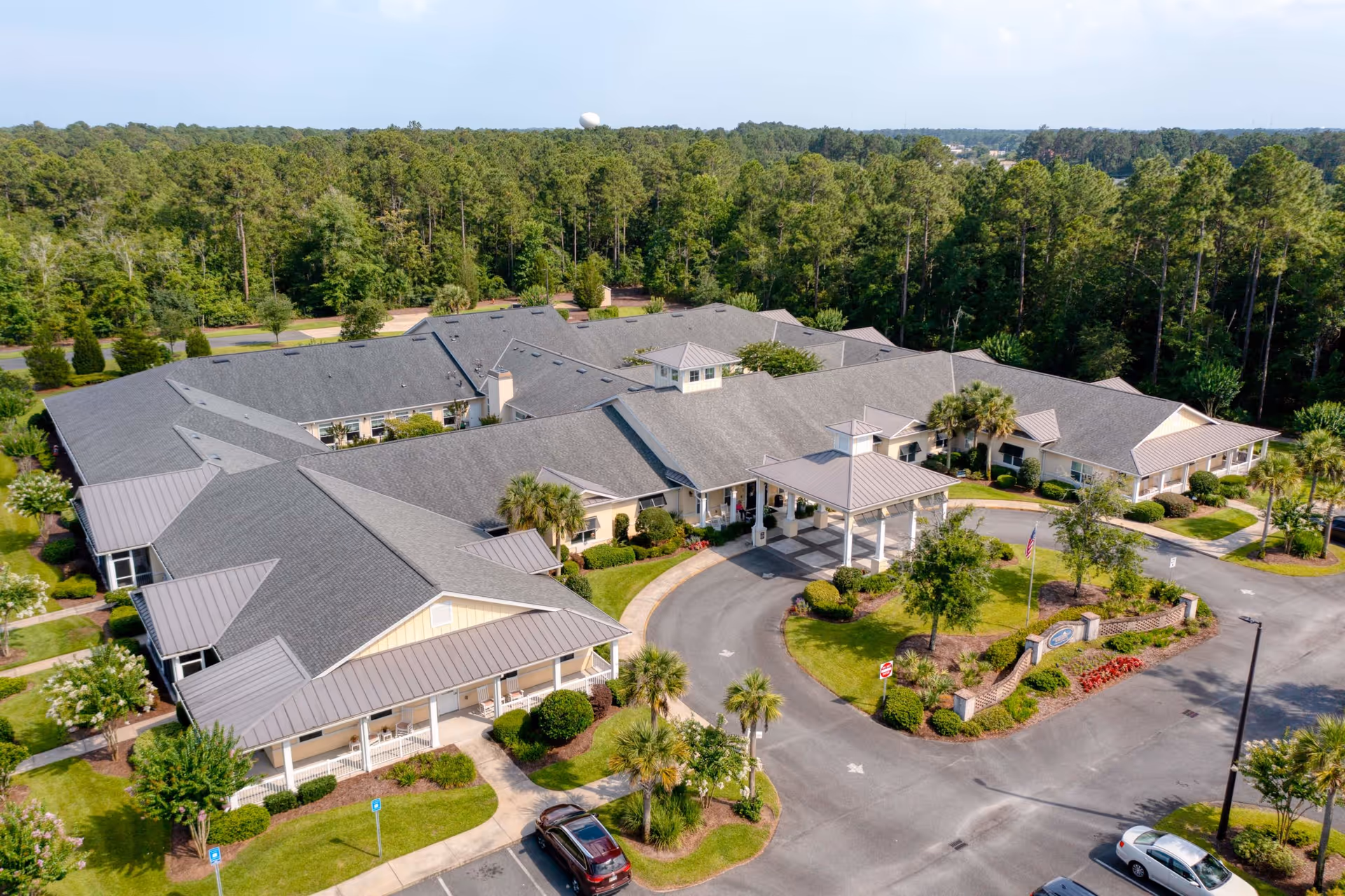 Aerial view of Addington Place of Brunswick, a single-story senior living facility surrounded by trees. The building has a large covered entrance with a circular driveway, landscaped gardens with palm trees and shrubs, and a parking area with several cars.
