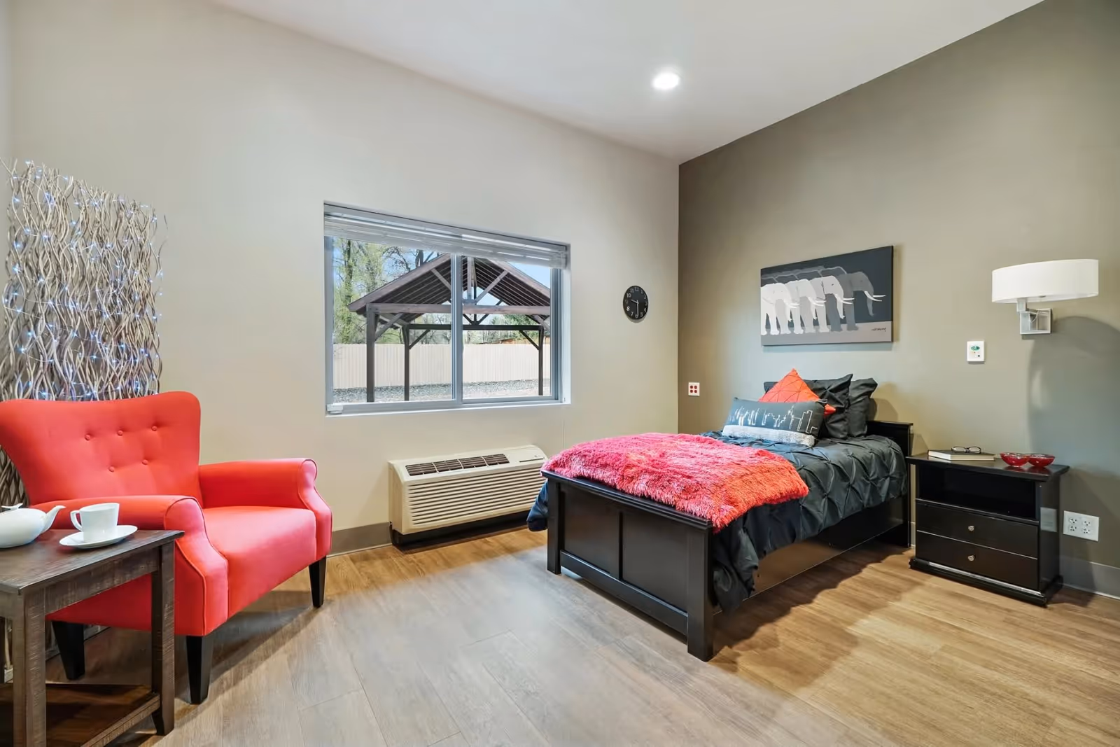 A cozy bedroom with a single black bed featuring black and red bedding, a red throw blanket, and decorative pillows. Next to the bed is a black nightstand with a book and two red bowls. A modern wall lamp is mounted above the nightstand. On the left side of the room, there is a bright red armchair next to a small wooden side table with a white teapot and cup. The room has light wood flooring, a large window showing an outdoor gazebo, and a wall clock. A decorative piece with lights is placed behind the armchair.