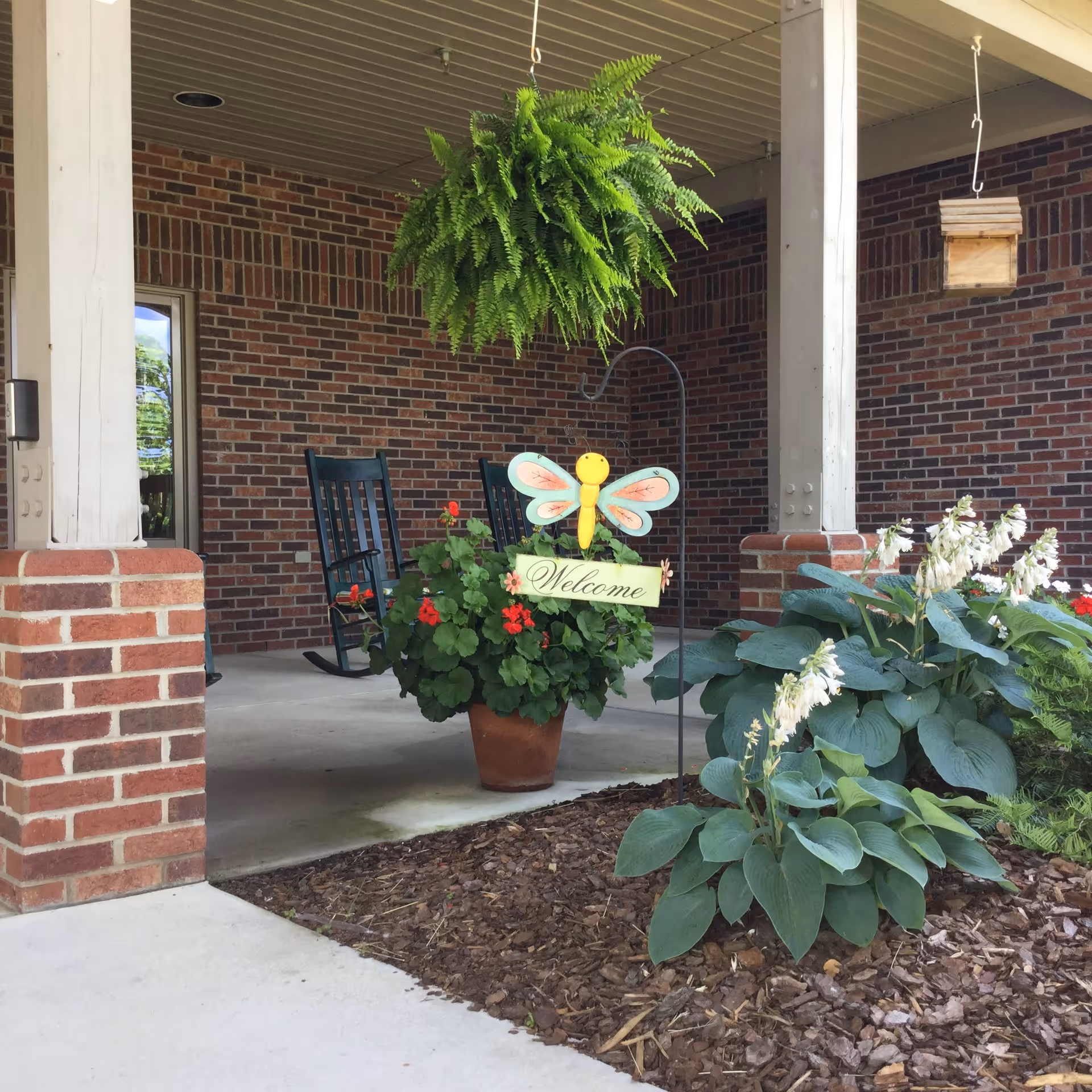 Covered brick front porch with potted plants, a hanging fern, rocking chairs, and a 'Welcome' sign.